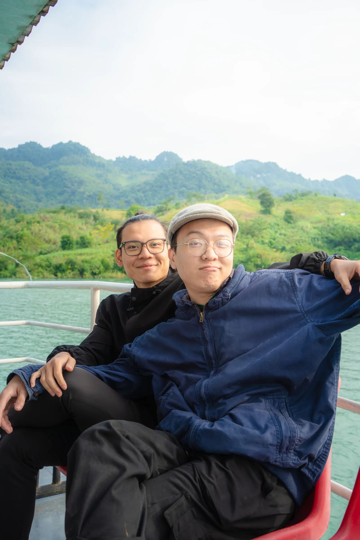 Hai người bạn ngồi cạnh nhau trên thuyền, phía sau là núi và hồ